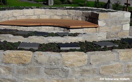 Holzbank Urnengräber Runder Steinsockel mit Holzbank und Pflanzenbepflanzung.