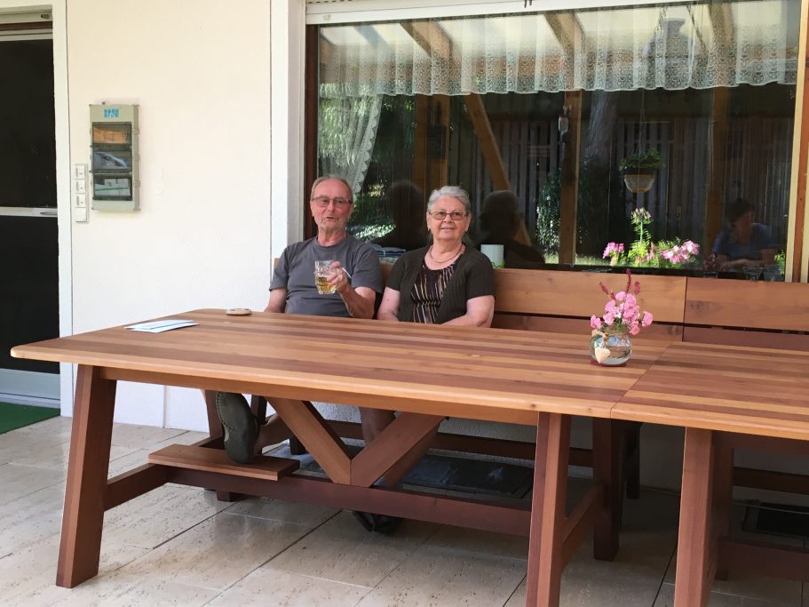 Terrassenmöbel Paar sitzt an einem großen Holztisch in einem hellen Raum mit Pflanzen und Dekoration.